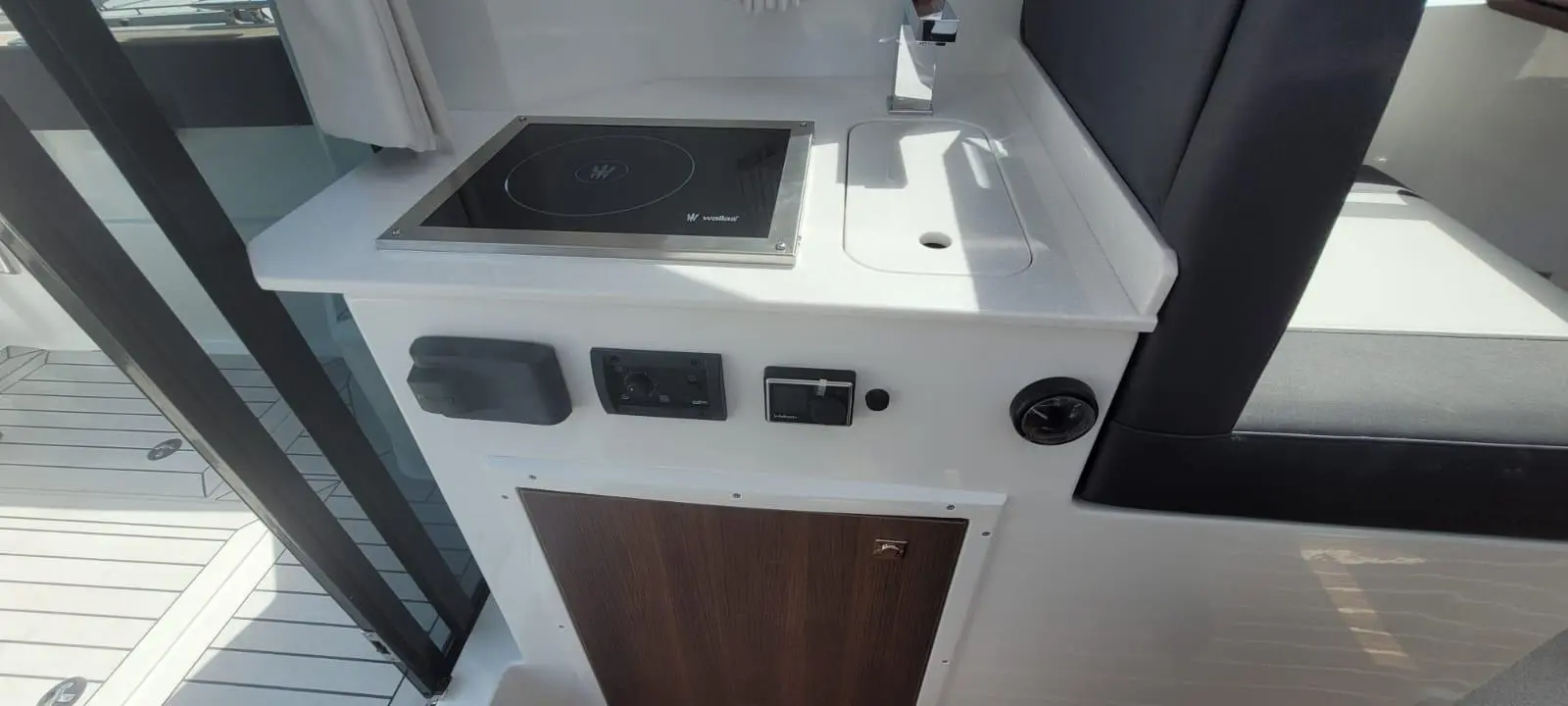 Boat kitchen area featuring an induction cooker, control panel, and wooden cabinet door. Sunlight illuminates the space.
